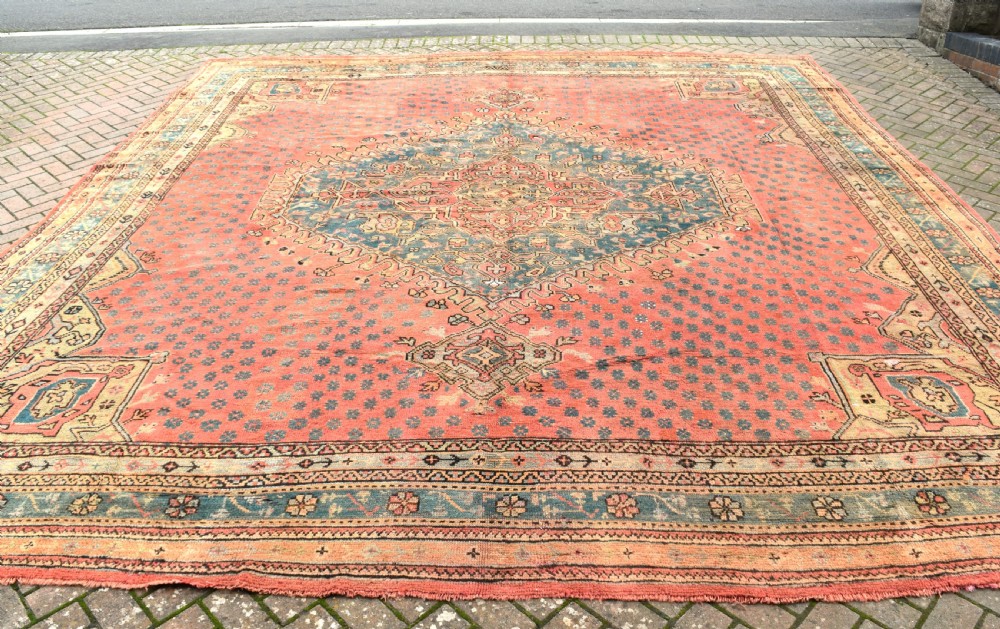 A large square hand-knotted wool Ushak carpet featuring a coral red field, a central teal medallion, cream corner spandrels, and patterned borders.