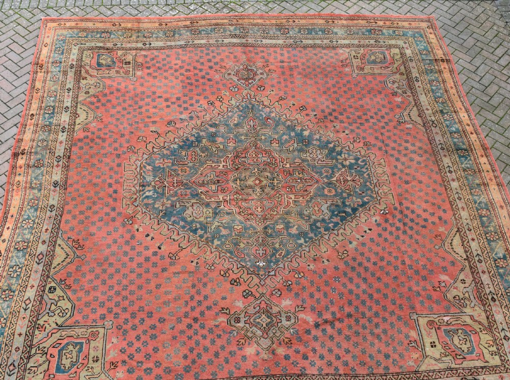 A large square hand-knotted wool Ushak carpet featuring a coral red field, a central teal medallion, cream corner spandrels, and patterned borders. View III