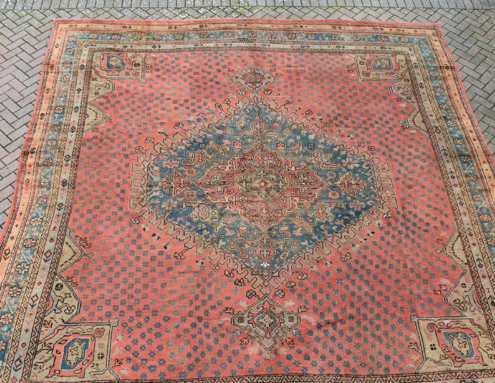 A large square hand-knotted wool Ushak carpet featuring a coral red field, a central teal medallion, cream corner spandrels, and patterned borders. View IV