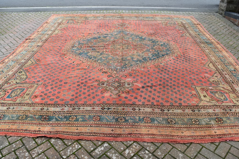 A large square hand-knotted wool Ushak carpet featuring a coral red field, a central teal medallion, cream corner spandrels, and patterned borders. View V