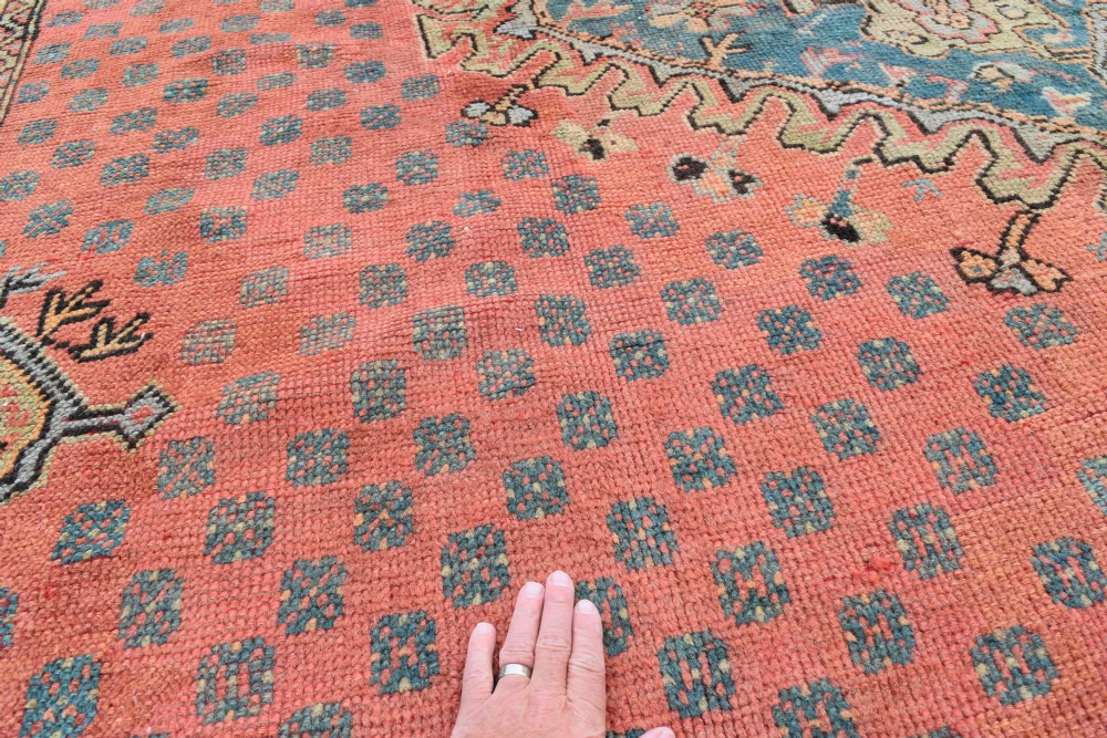 A large square hand-knotted wool Ushak carpet featuring a coral red field, a central teal medallion, cream corner spandrels, and patterned borders. View VII