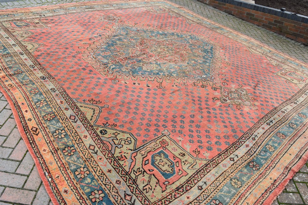 A large square hand-knotted wool Ushak carpet featuring a coral red field, a central teal medallion, cream corner spandrels, and patterned borders. View IX