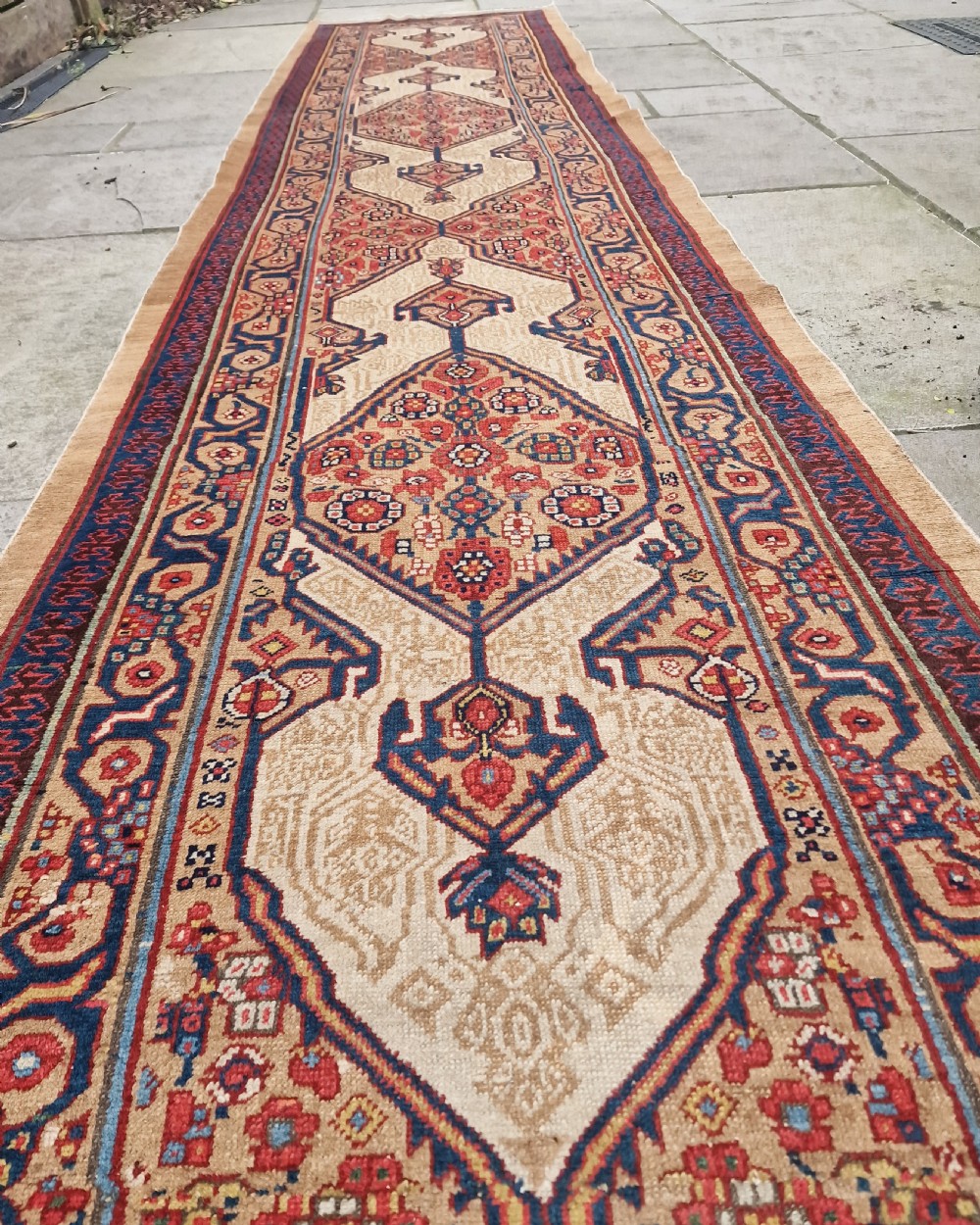 A long, narrow antique Persian Serab wool carpet runner featuring geometric diamond medallions on a camel-coloured field with red and navy patterned borders. View II