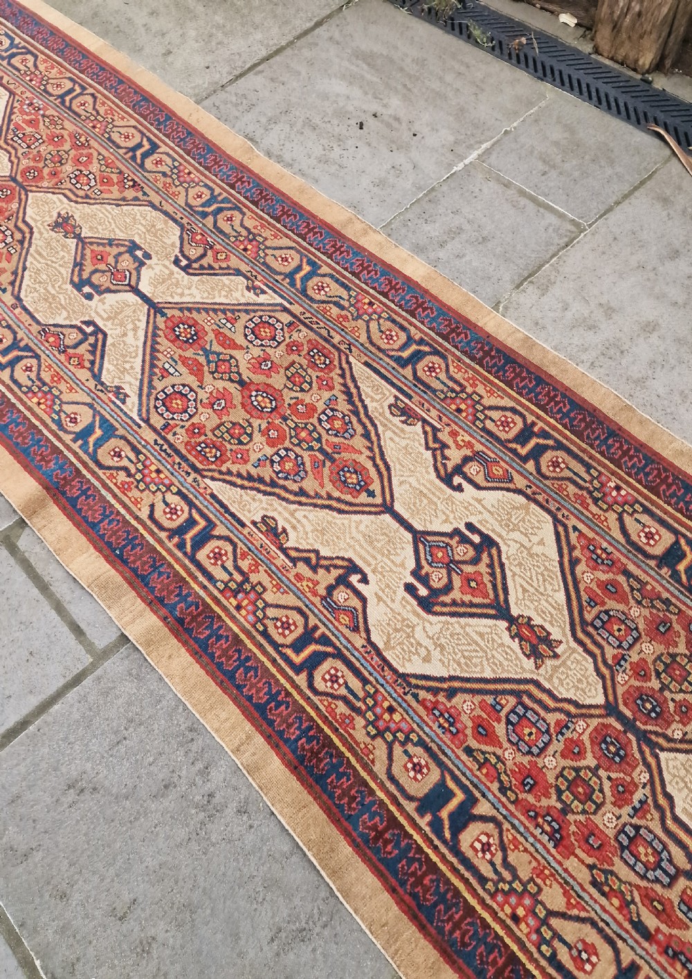 A long, narrow antique Persian Serab wool carpet runner featuring geometric diamond medallions on a camel-coloured field with red and navy patterned borders. View VI