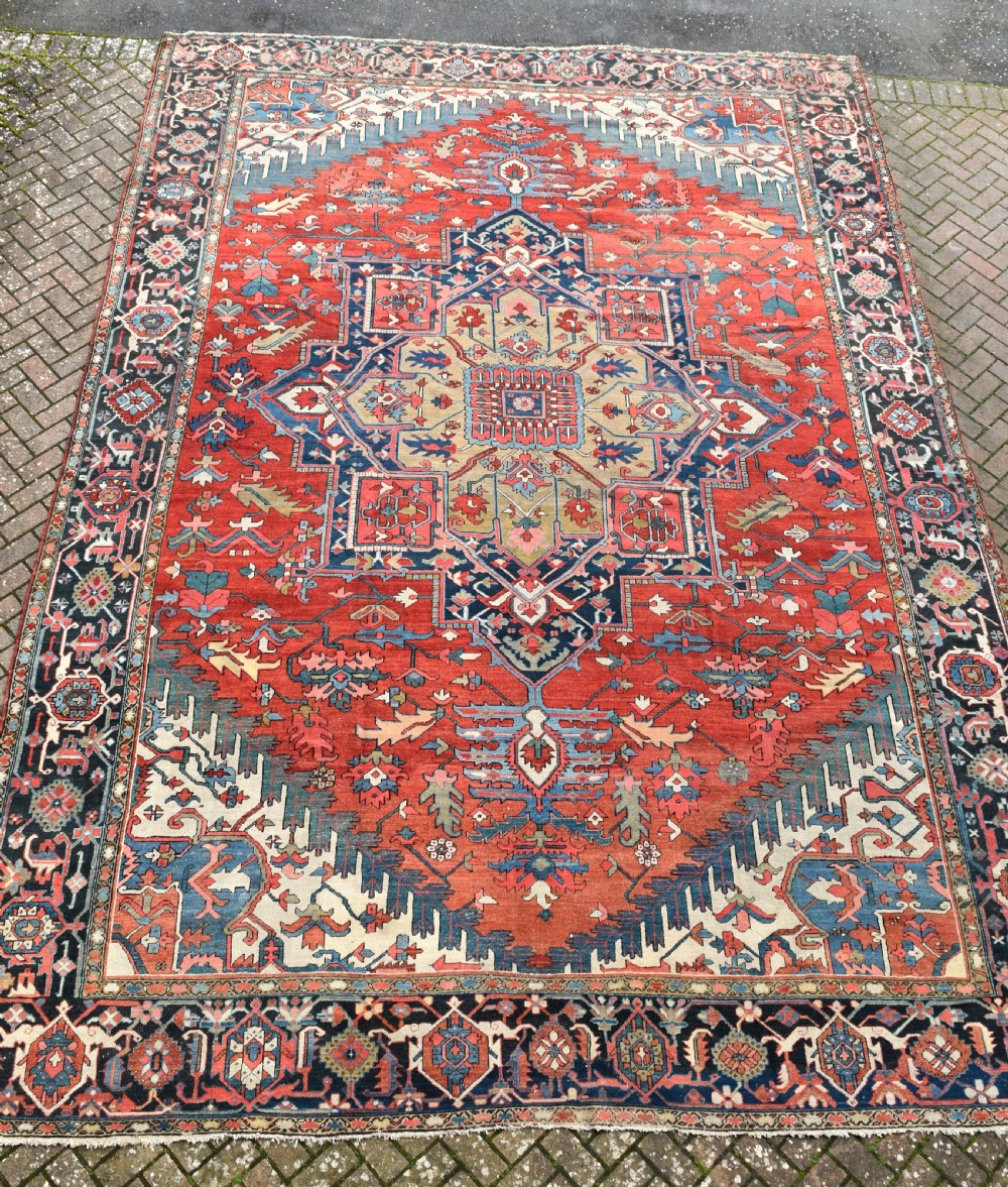 An oversize antique Heriz wool carpet featuring a prominent geometric central medallion in navy blue, beige, and red on a terracotta field with patterned spandrels and border.