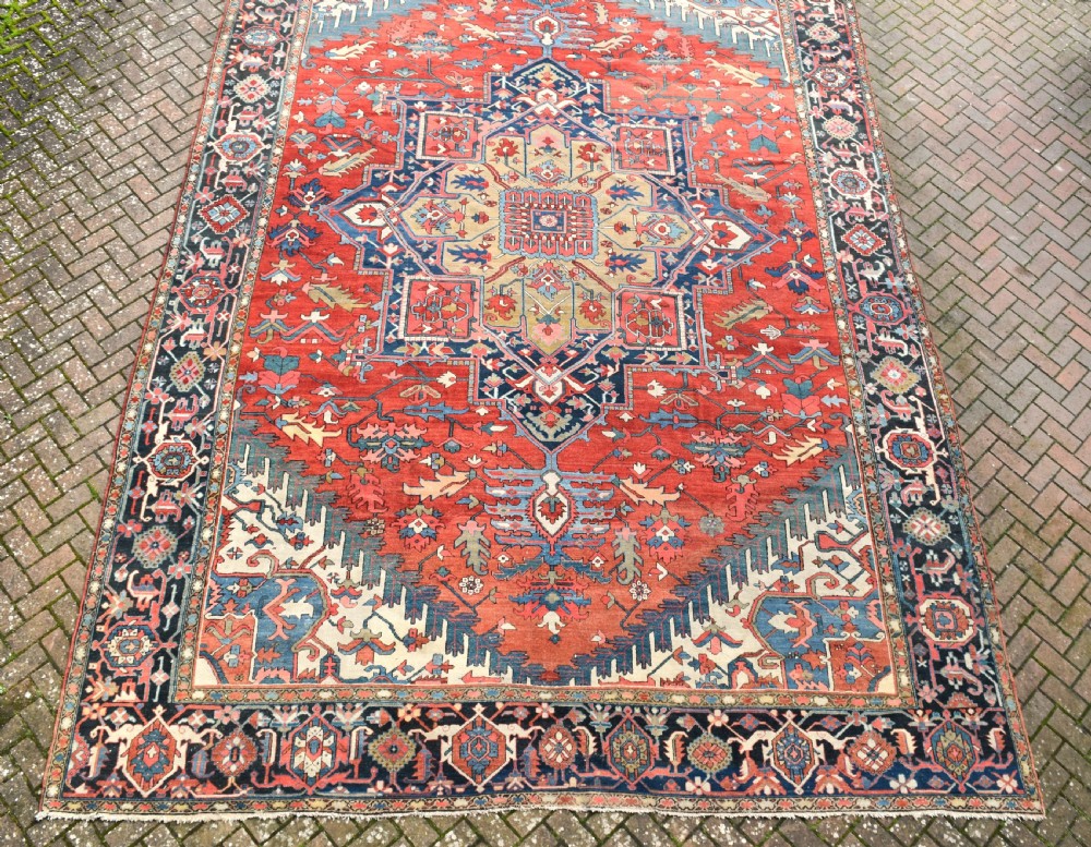 An oversize antique Heriz wool carpet featuring a prominent geometric central medallion in navy blue, beige, and red on a terracotta field with patterned spandrels and border. View II