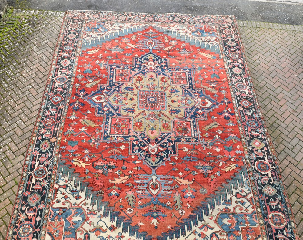 An oversize antique Heriz wool carpet featuring a prominent geometric central medallion in navy blue, beige, and red on a terracotta field with patterned spandrels and border. View III