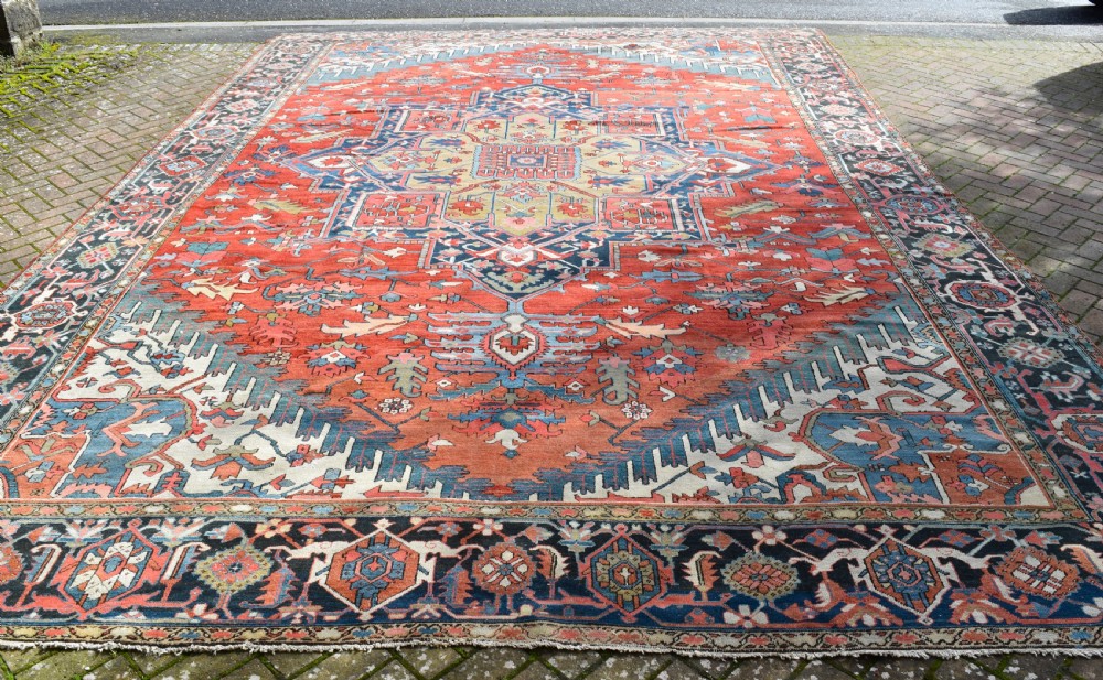 An oversize antique Heriz wool carpet featuring a prominent geometric central medallion in navy blue, beige, and red on a terracotta field with patterned spandrels and border. View IV