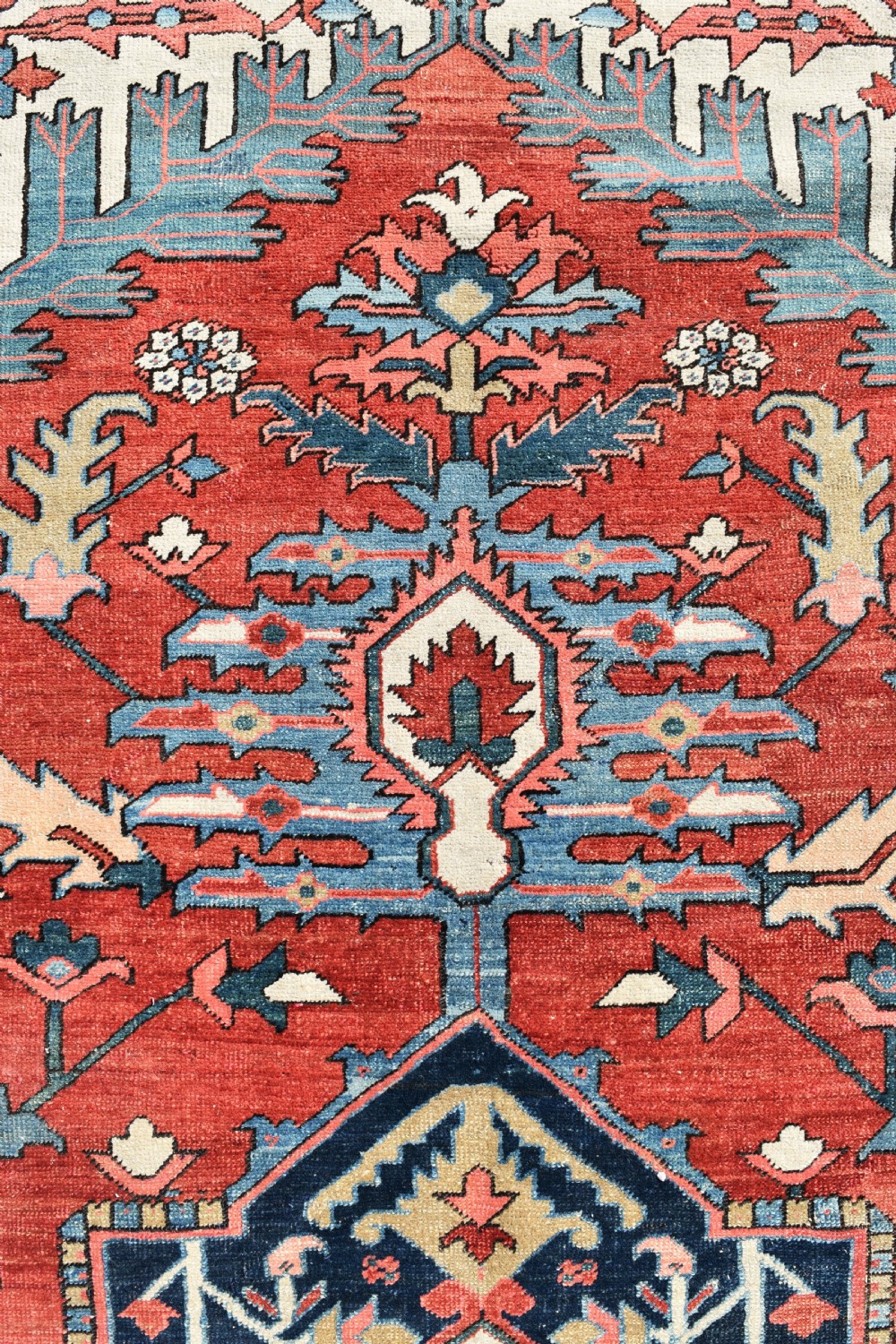 An oversize antique Heriz wool carpet featuring a prominent geometric central medallion in navy blue, beige, and red on a terracotta field with patterned spandrels and border. View V