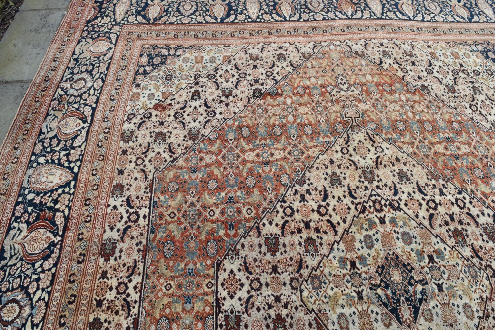 A large antique hand-knotted Tabriz carpet featuring an intricate central medallion in terracotta and cream framed by a wide navy floral border. View IV