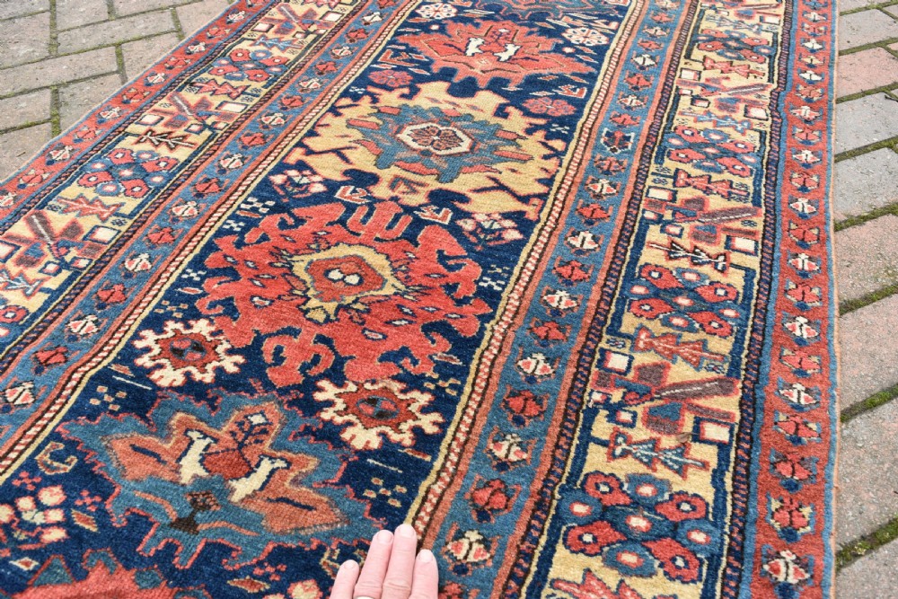 A long hand-knotted wool runner rug featuring repeating geometric medallions in red, navy blue, and yellow along framed patterned borders. View VI