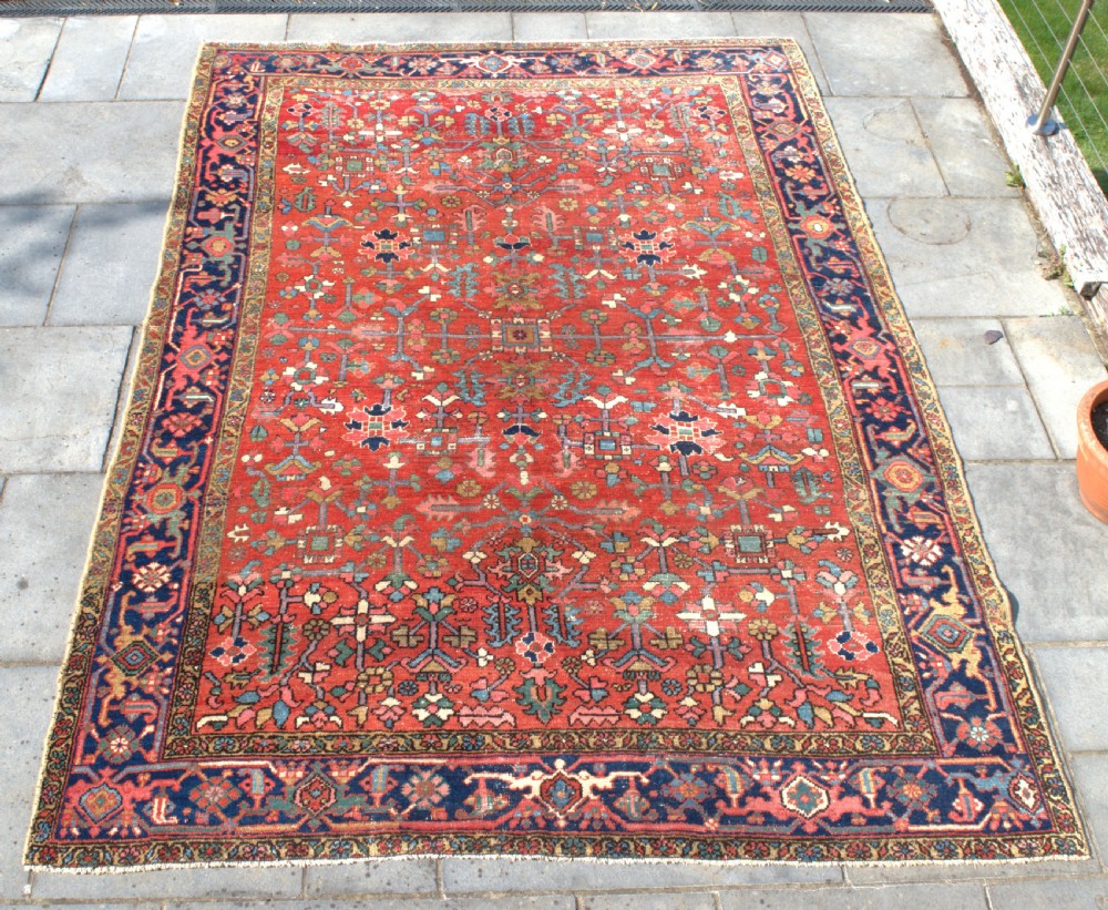 A large antique Persian Heriz wool carpet featuring a terracotta red field with geometric floral motifs and a dark navy blue outer border.