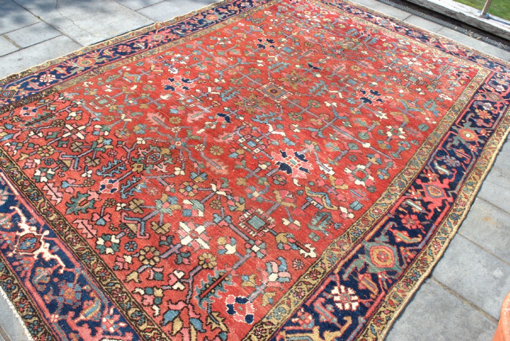 A large antique Persian Heriz wool carpet featuring a terracotta red field with geometric floral motifs and a dark navy blue outer border. View III