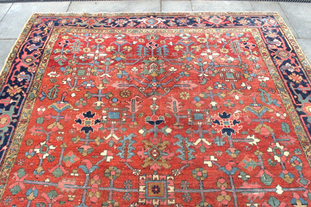 A large antique Persian Heriz wool carpet featuring a terracotta red field with geometric floral motifs and a dark navy blue outer border. View IV