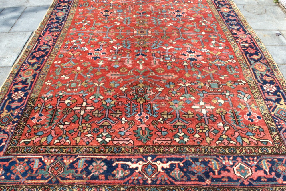 A large antique Persian Heriz wool carpet featuring a terracotta red field with geometric floral motifs and a dark navy blue outer border. View V