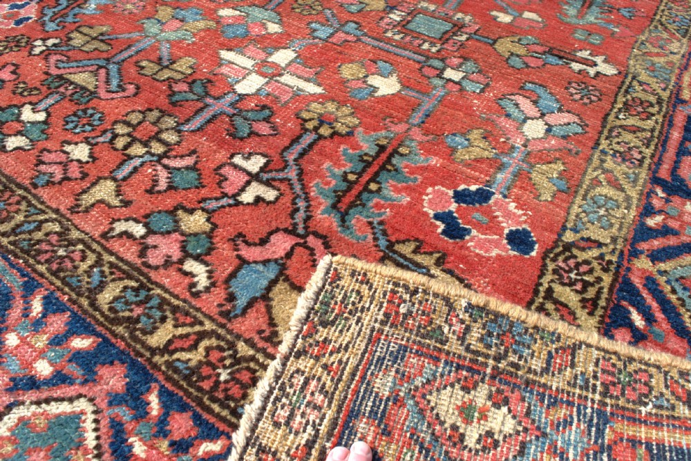 A large antique Persian Heriz wool carpet featuring a terracotta red field with geometric floral motifs and a dark navy blue outer border. View VI