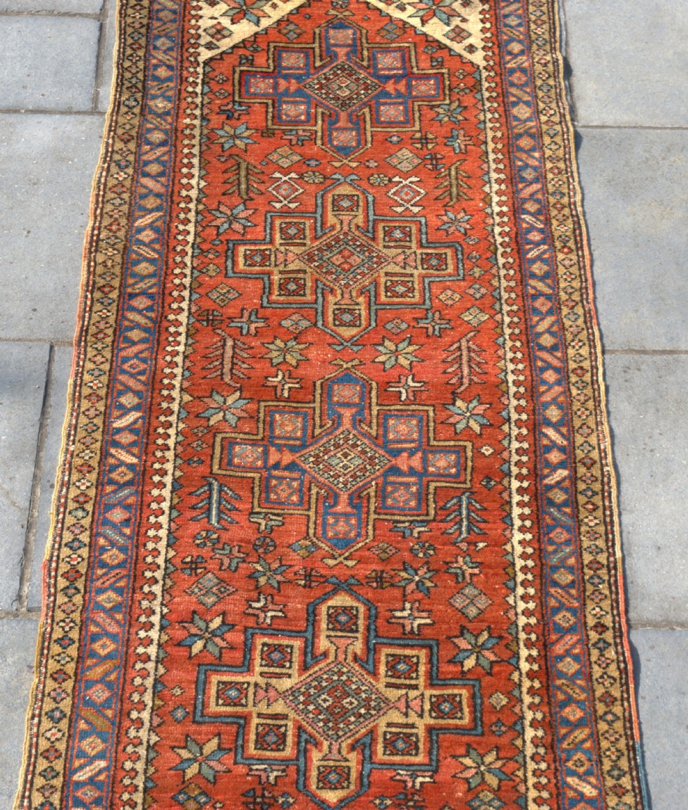 An antique Persian Heriz wool runner rug featuring repeating geometric medallions on a terracotta red field with blue and cream borders. View III