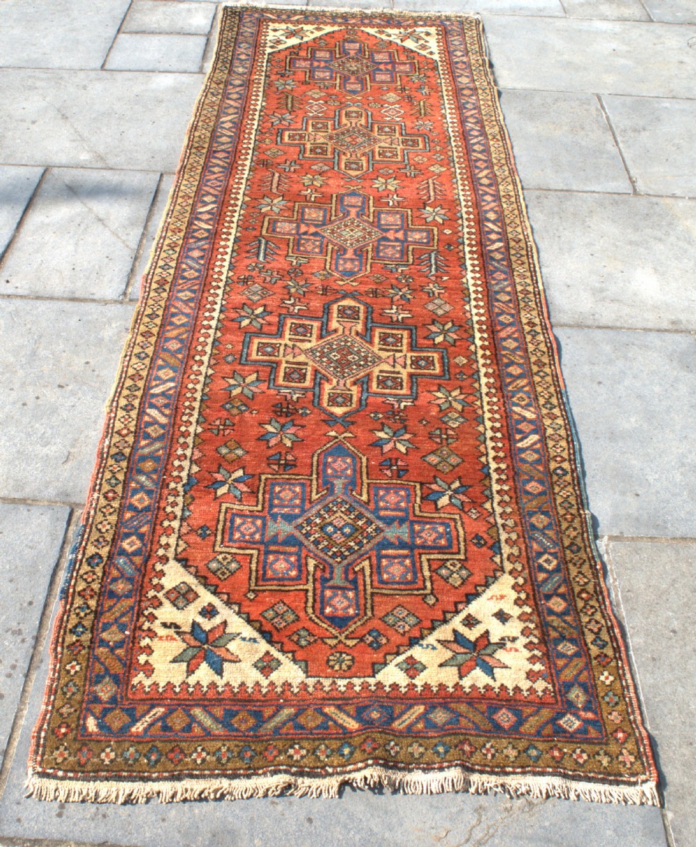 An antique Persian Heriz wool runner rug featuring repeating geometric medallions on a terracotta red field with blue and cream borders. View V