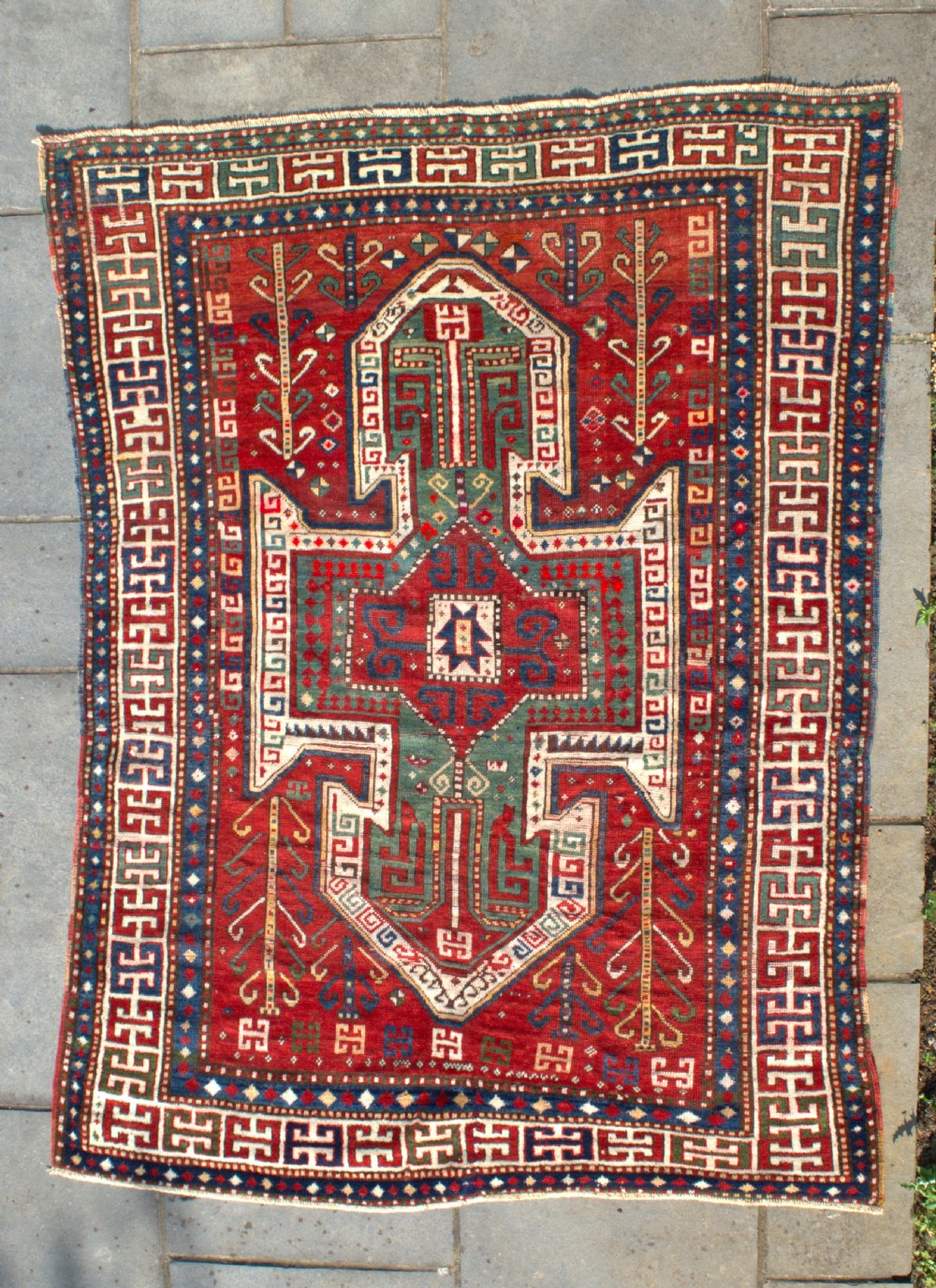 An antique hand-knotted Caucasian Sewan Kazak wool rug featuring a green central shield motif on a red ground with geometric borders.