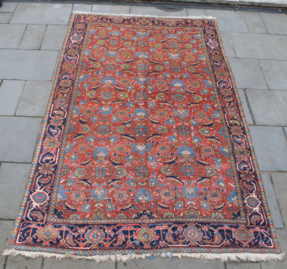 An antique hand-knotted Persian Heriz wool carpet featuring a dense geometric floral pattern on a rust red field with a dark blue ornamental border.