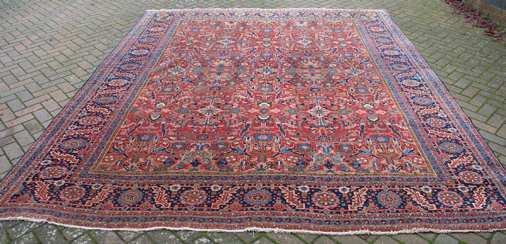 A large hand-knotted wool Heriz carpet featuring an intricate red central field decorated with geometric floral motifs and a dark navy border.