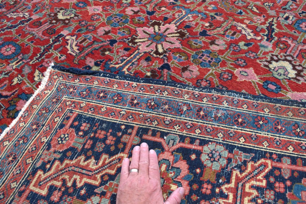 A large hand-knotted wool Heriz carpet featuring an intricate red central field decorated with geometric floral motifs and a dark navy border. View IV
