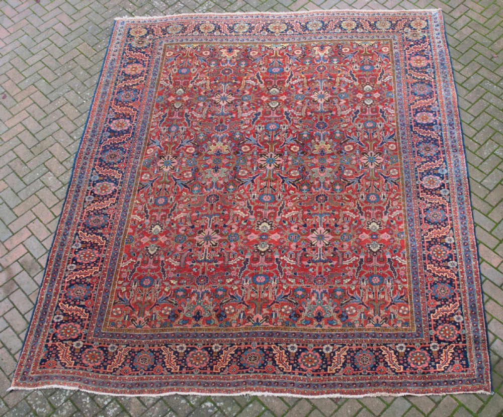 A large hand-knotted wool Heriz carpet featuring an intricate red central field decorated with geometric floral motifs and a dark navy border. View V