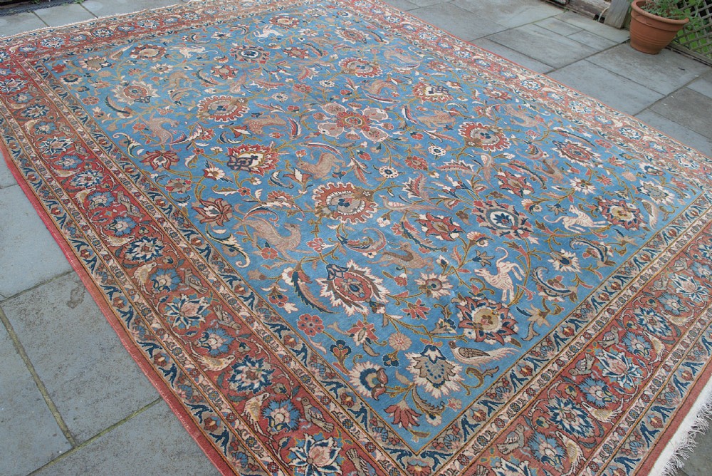 A large hand-knotted wool Veramin carpet featuring a central sky-blue field detailed with animals, birds, and floral vine scrolls inside a red multi-bordered frame. View VI