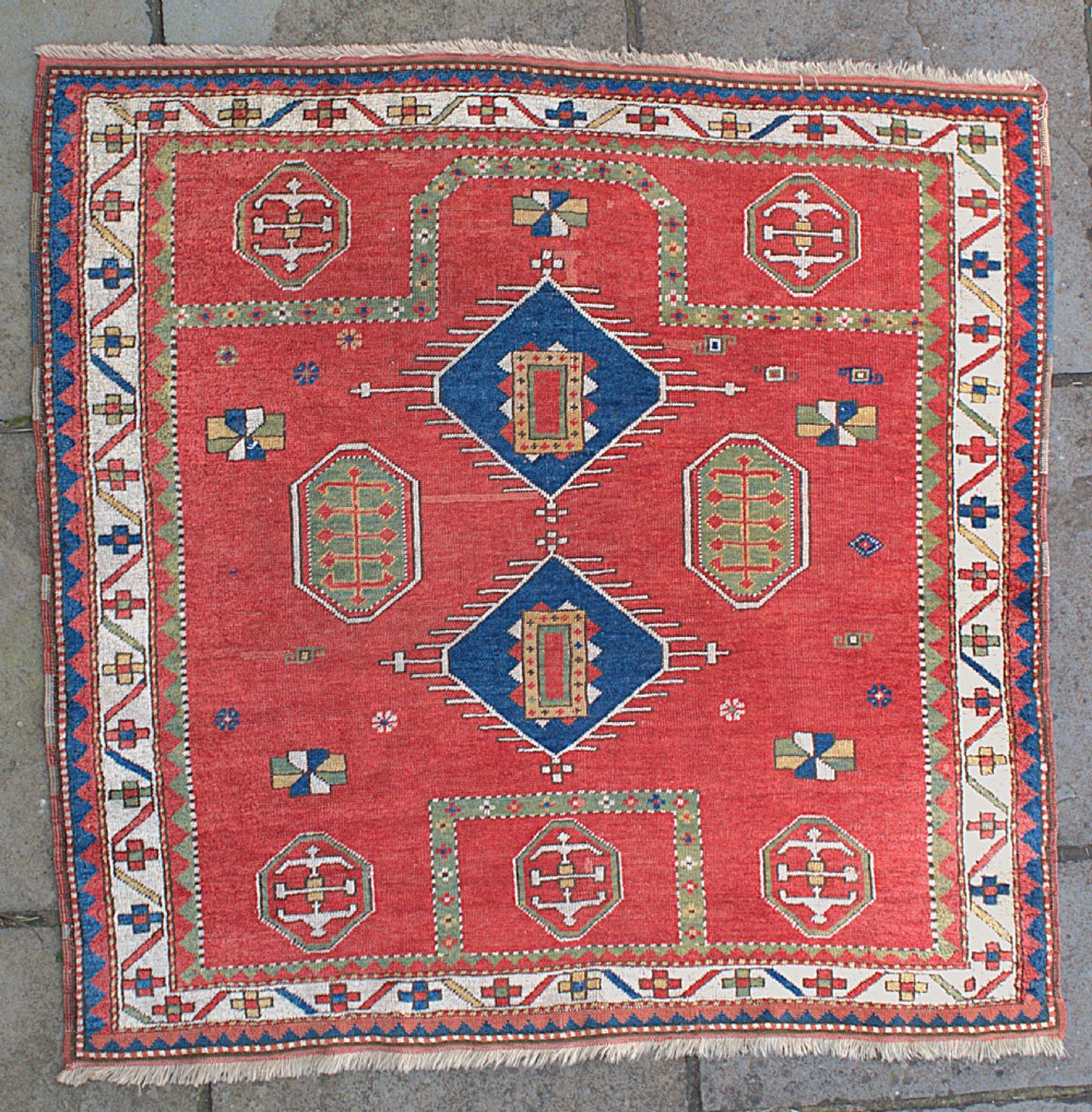 An antique square Kazak wool prayer rug featuring a red ground, central blue diamond medallions, geometric octagonal motifs, and a patterned cream border.