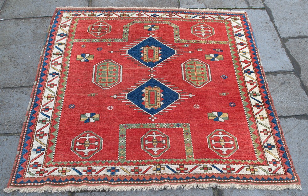 An antique square Kazak wool prayer rug featuring a red ground, central blue diamond medallions, geometric octagonal motifs, and a patterned cream border. View II