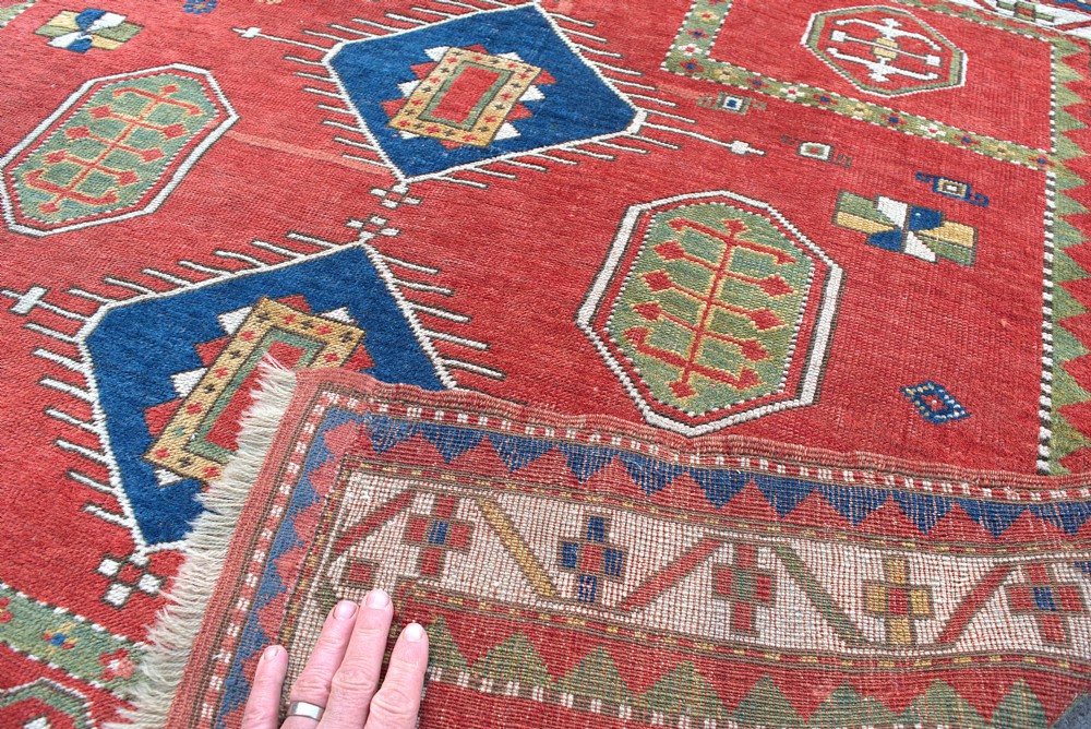 An antique square Kazak wool prayer rug featuring a red ground, central blue diamond medallions, geometric octagonal motifs, and a patterned cream border. View III