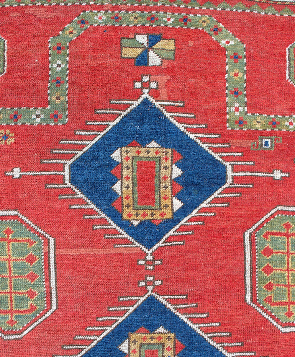An antique square Kazak wool prayer rug featuring a red ground, central blue diamond medallions, geometric octagonal motifs, and a patterned cream border. View IV