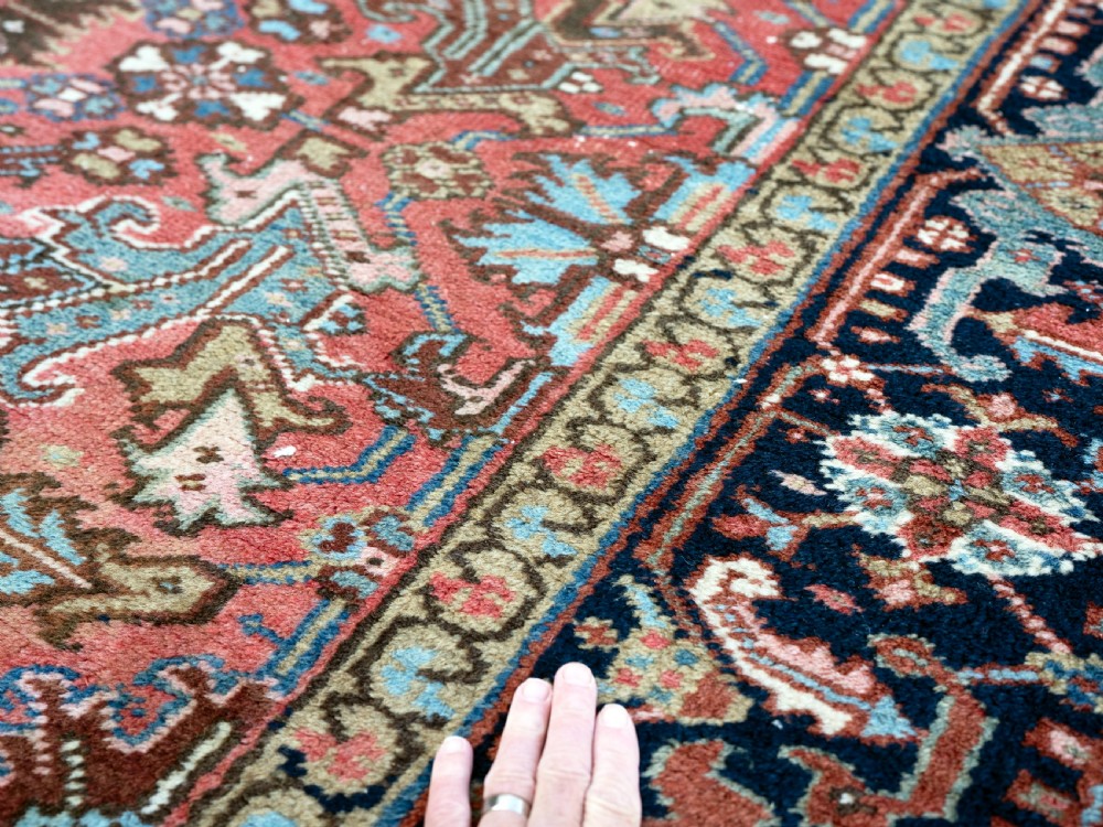An antique Heriz wool carpet featuring a rust red field with geometric floral patterns framed by a dark navy patterned border. View IV