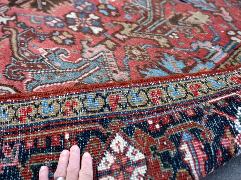 An antique Heriz wool carpet featuring a rust red field with geometric floral patterns framed by a dark navy patterned border. View V