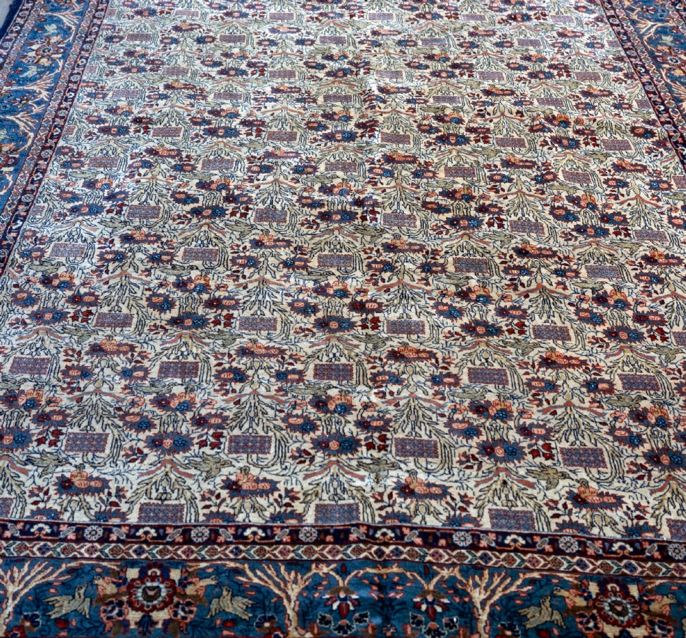A rectangular hand-knotted Persian Qum carpet featuring an intricate botanical pattern on a cream field framed by a dark blue patterned border. View II