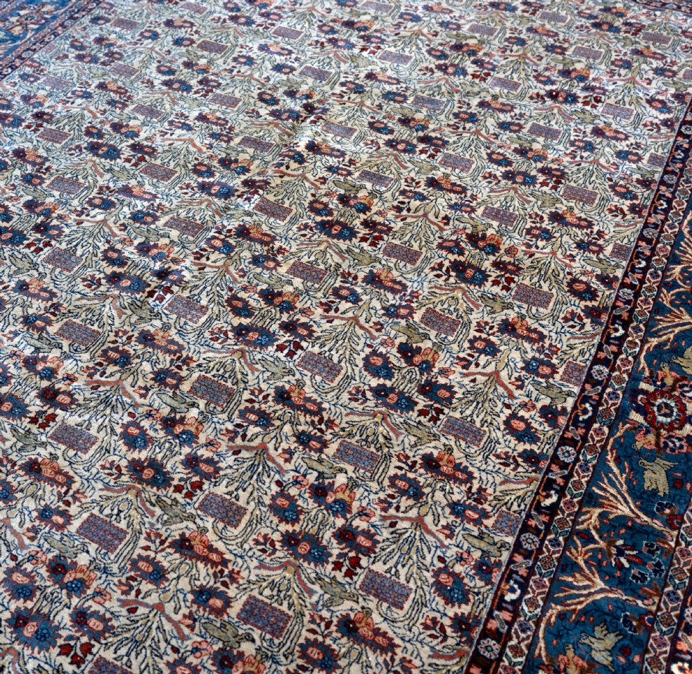 A rectangular hand-knotted Persian Qum carpet featuring an intricate botanical pattern on a cream field framed by a dark blue patterned border. View III