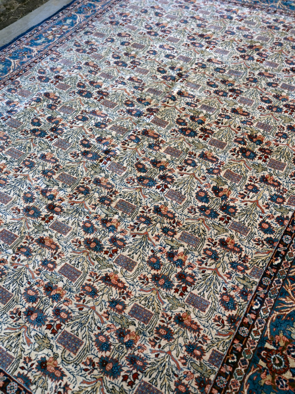 A rectangular hand-knotted Persian Qum carpet featuring an intricate botanical pattern on a cream field framed by a dark blue patterned border. View V