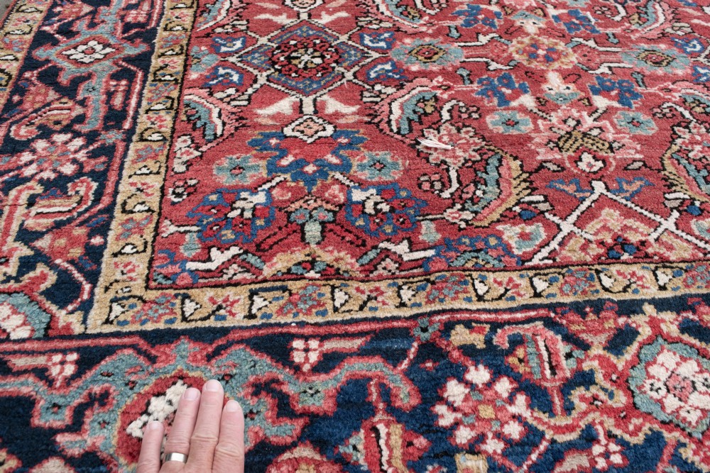 An antique Persian Heriz wool rug featuring a rich red field decorated with intricate geometric floral motifs framed by a dark navy blue patterned border. View II