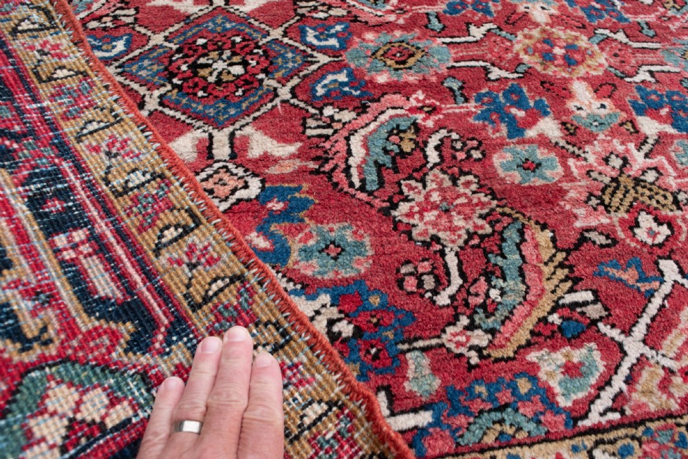 An antique Persian Heriz wool rug featuring a rich red field decorated with intricate geometric floral motifs framed by a dark navy blue patterned border. View III