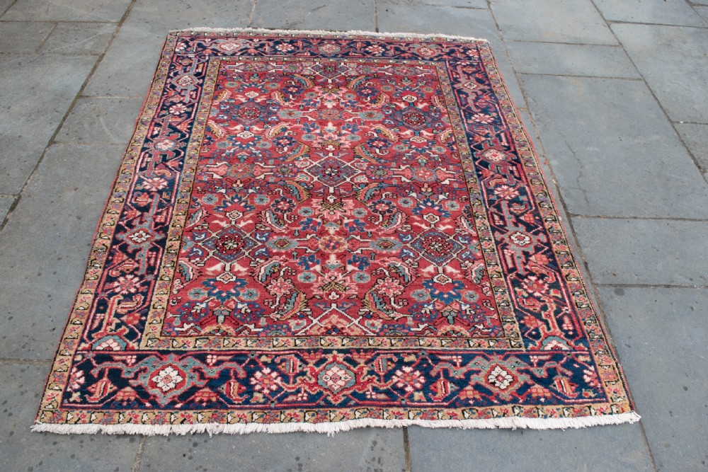 An antique Persian Heriz wool rug featuring a rich red field decorated with intricate geometric floral motifs framed by a dark navy blue patterned border. View IV