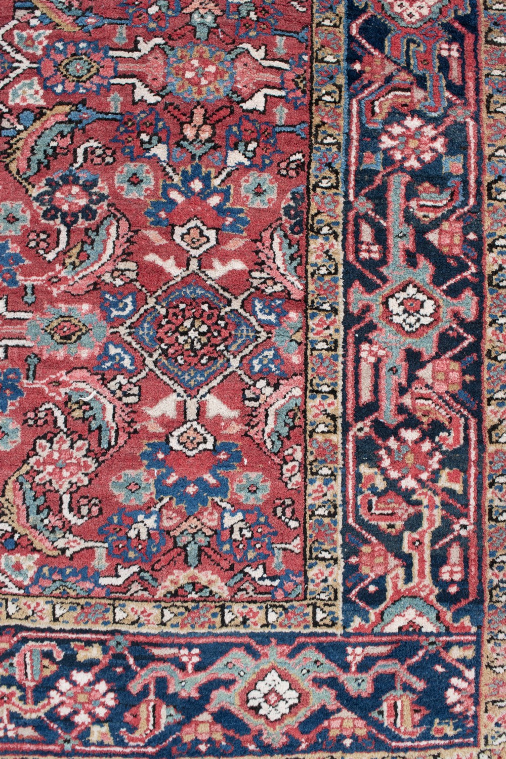 An antique Persian Heriz wool rug featuring a rich red field decorated with intricate geometric floral motifs framed by a dark navy blue patterned border. View V