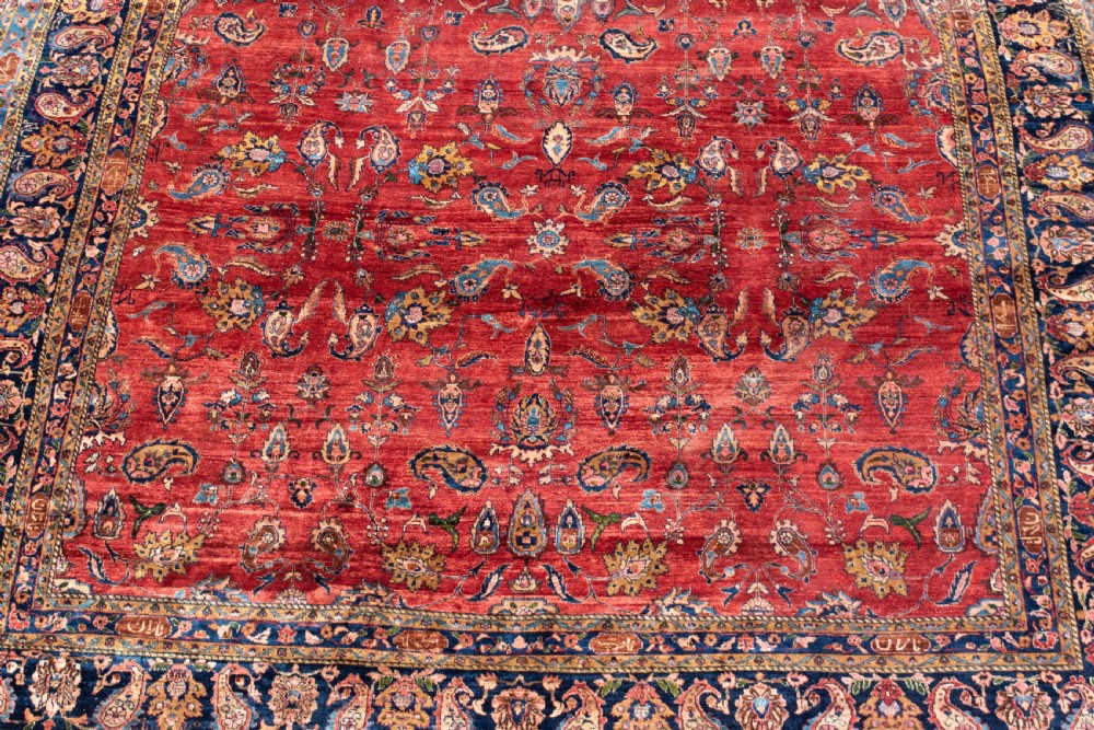 A large hand-knotted Persian Kashan carpet featuring a detailed red field covered in floral motifs framed by a multi-tiered navy blue border. View IV