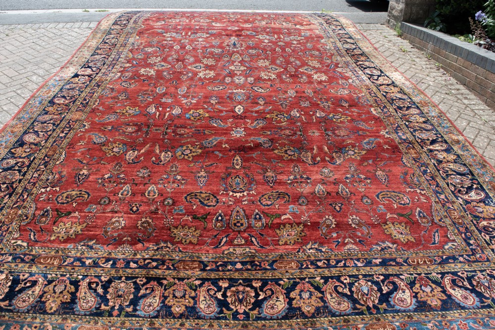 A large hand-knotted Persian Kashan carpet featuring a detailed red field covered in floral motifs framed by a multi-tiered navy blue border. View V