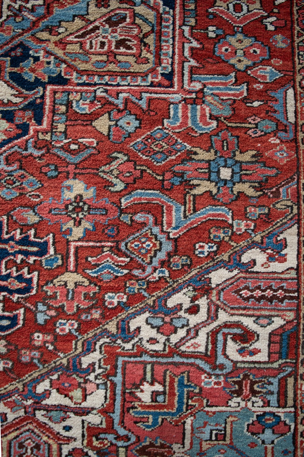 A large hand-knotted wool Heriz carpet featuring a central geometric medallion framed by intricate borders in shades of rust red, navy blue, and ivory. View III