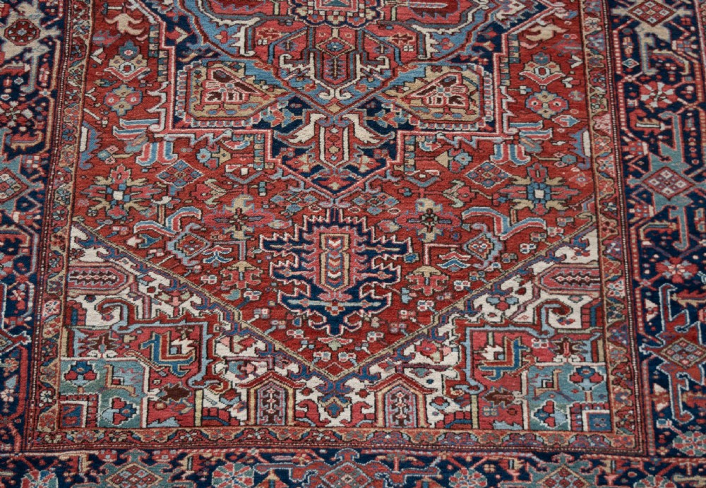 A large hand-knotted wool Heriz carpet featuring a central geometric medallion framed by intricate borders in shades of rust red, navy blue, and ivory. View V