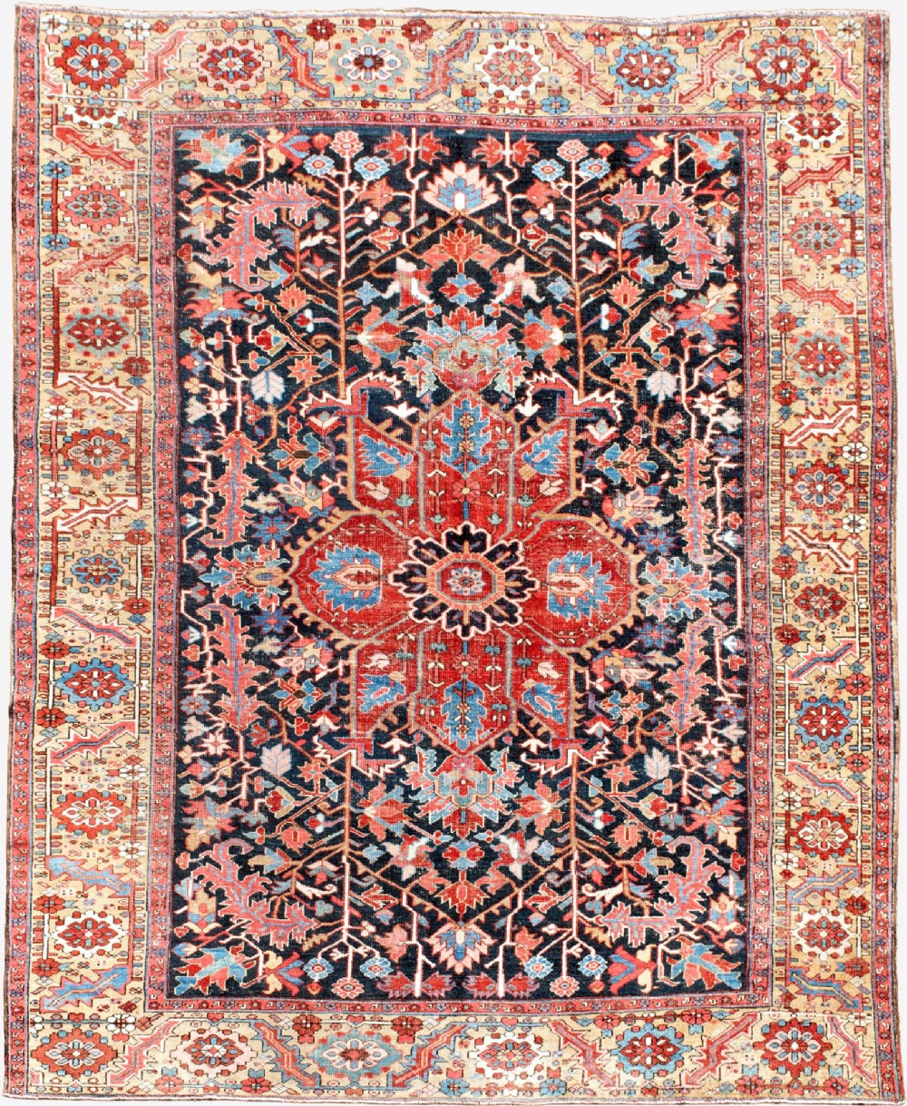 An antique Heriz wool carpet featuring a large red central geometric medallion on a dark navy blue field surrounded by an elaborate cream motif border.