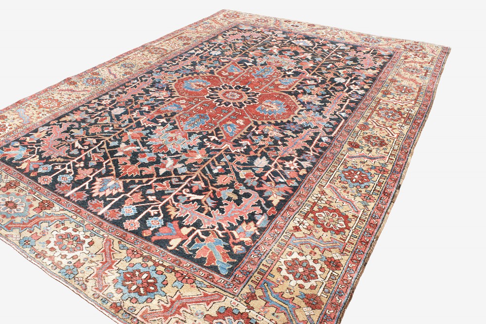 An antique Heriz wool carpet featuring a large red central geometric medallion on a dark navy blue field surrounded by an elaborate cream motif border. View V