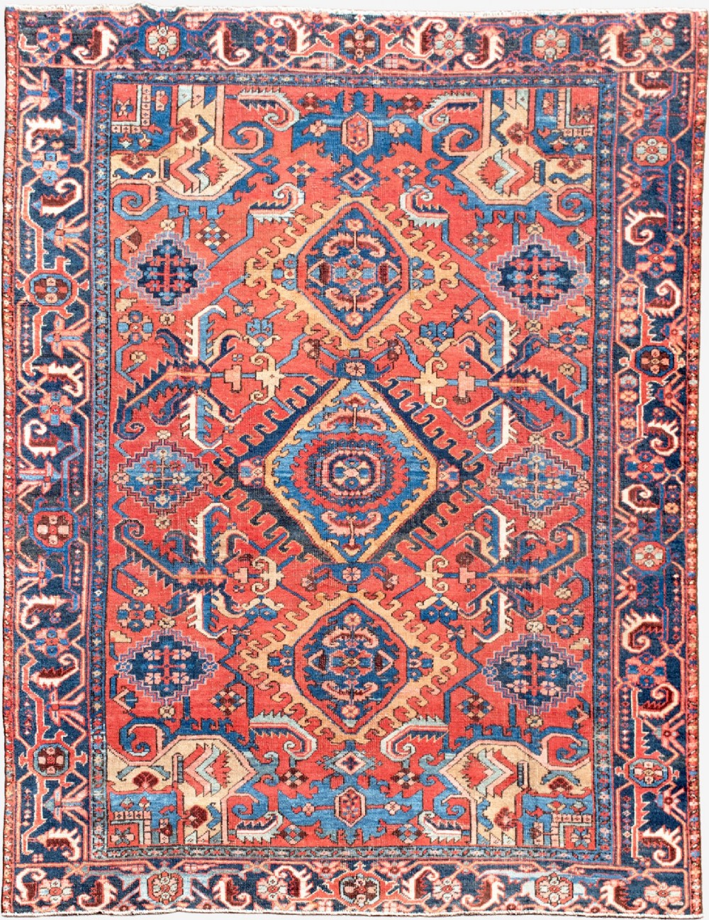 An antique Heriz wool carpet featuring a prominent geometric central medallion in blue and terracotta surrounded by angular botanical motifs and a patterned navy border.