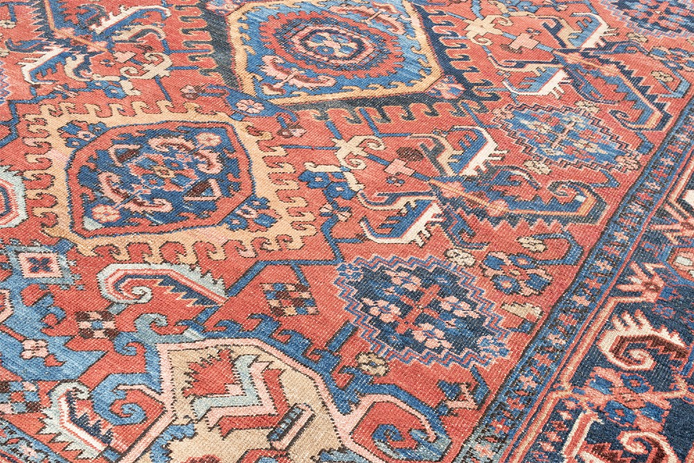 An antique Heriz wool carpet featuring a prominent geometric central medallion in blue and terracotta surrounded by angular botanical motifs and a patterned navy border. View V
