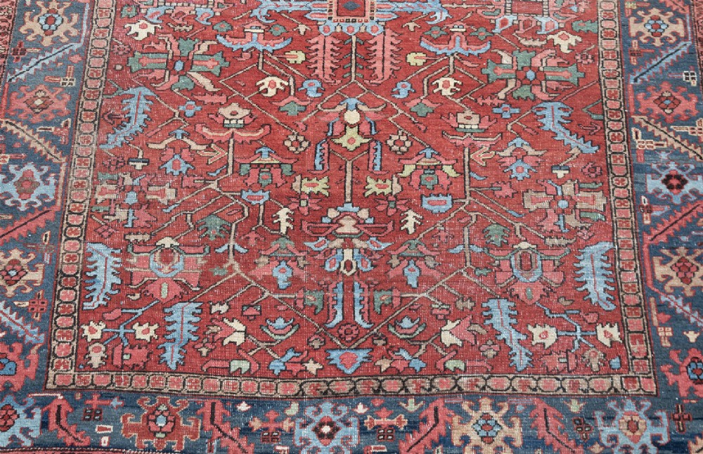 An antique hand-knotted Persian Heriz carpet featuring a terracotta red field with geometric motifs enclosed by a dark blue patterned border. View VI